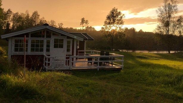 A cabin with a deck, surrounded by a grassy area and trees, with a lake in the background. - Charming 2-bed House at the Lake Close Gustavsfors (Gustavsfors)