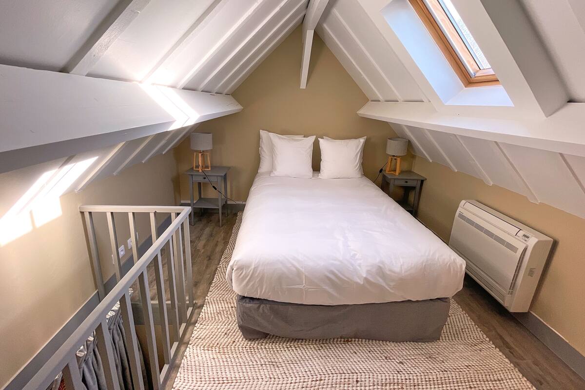 A bedroom with a sloped ceiling, a skylight, a bed with white bedding, two bedside tables with lamps, and a radiator.