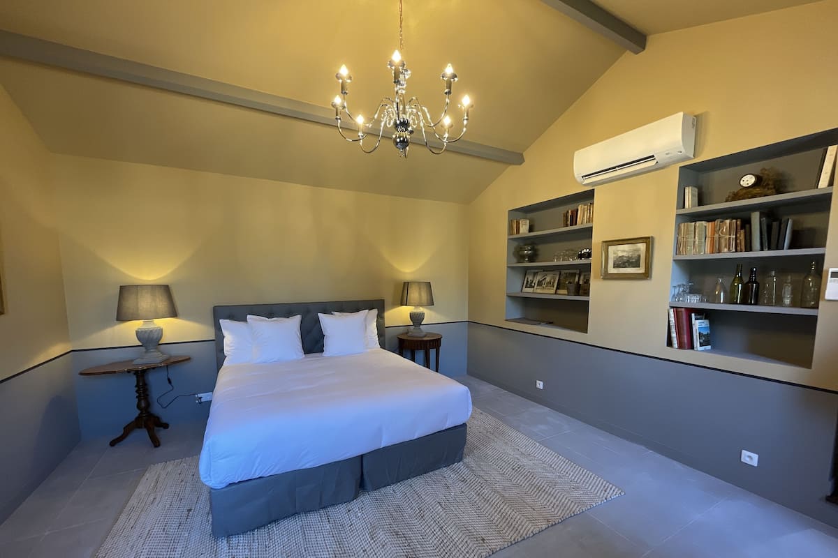A bedroom with a large bed, a chandelier, bedside lamps, a bookshelf, and an air conditioning unit.
