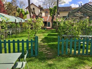 A courtyard with green tables and chairs, a brick pathway, and a building with a tiled roof in the background. - Le Fruit Défendu (Rueil-Malmaison)