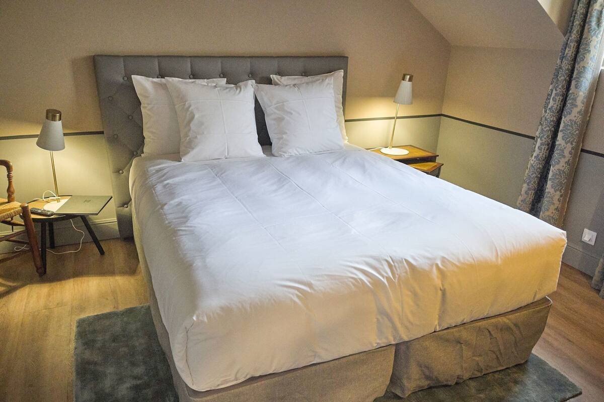 A neatly made bed with a grey headboard, two bedside lamps, a laptop on a side table, and a wooden chair.