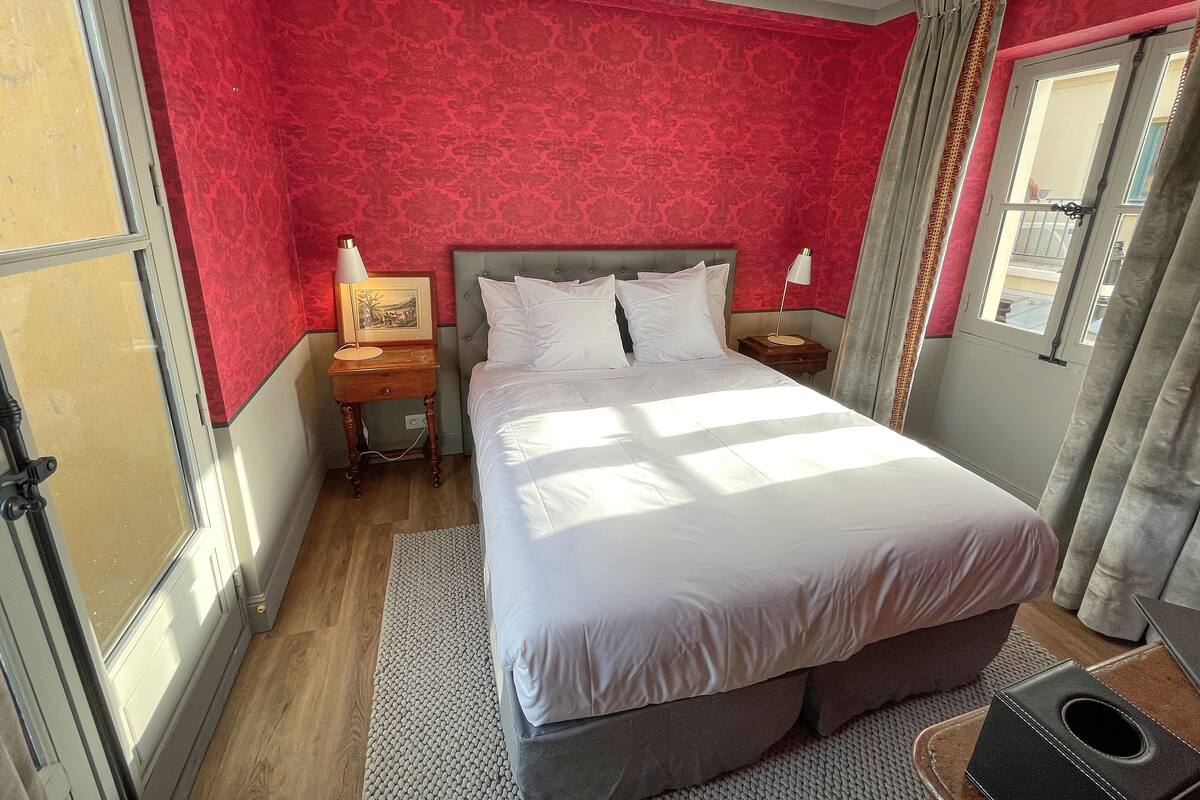 A bedroom with a large bed, a bedside table with a lamp, a framed picture, and a window with curtains.
