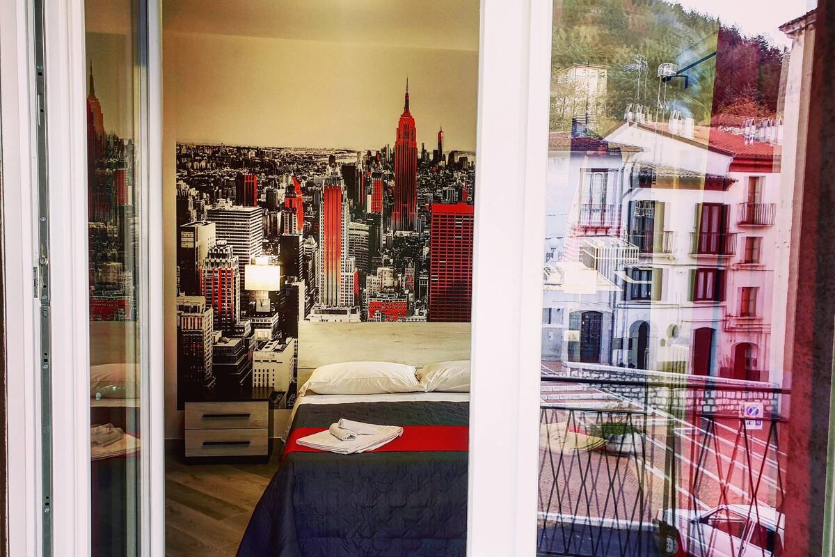 Une chambre avec une fresque représentant un paysage urbain sur le mur, un lit recouvert d’une courtepointe rouge et noire, et une vue sur un balcon avec une balustrade.