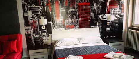 A hotel room with a cityscape mural on the wall, a bed with a red blanket, and a red chair.