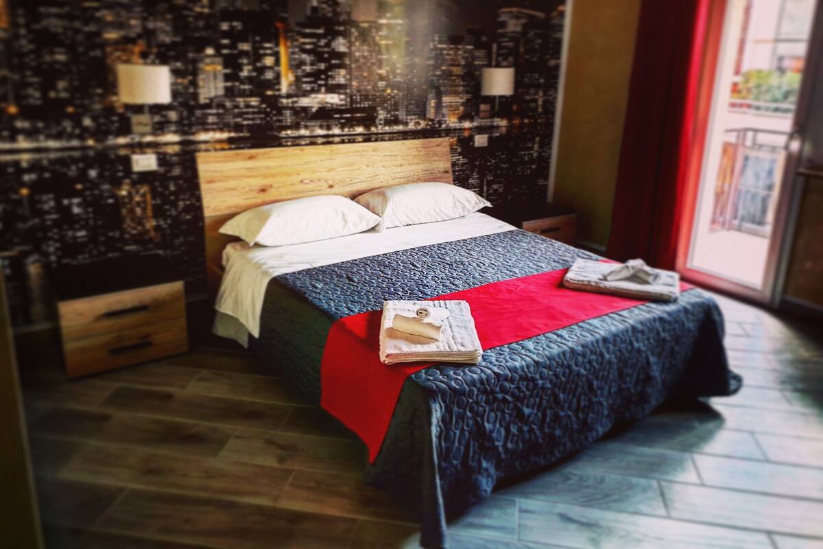 Un lit avec une tête de lit en bois et un mur décoré d’un motif représentant un paysage urbain, ainsi qu’une couverture rouge.