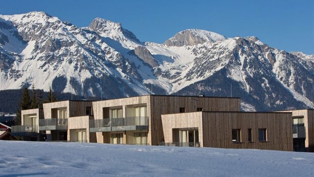 Un edificio moderno con ampie finestre e balconi, sullo sfondo di montagne innevate.