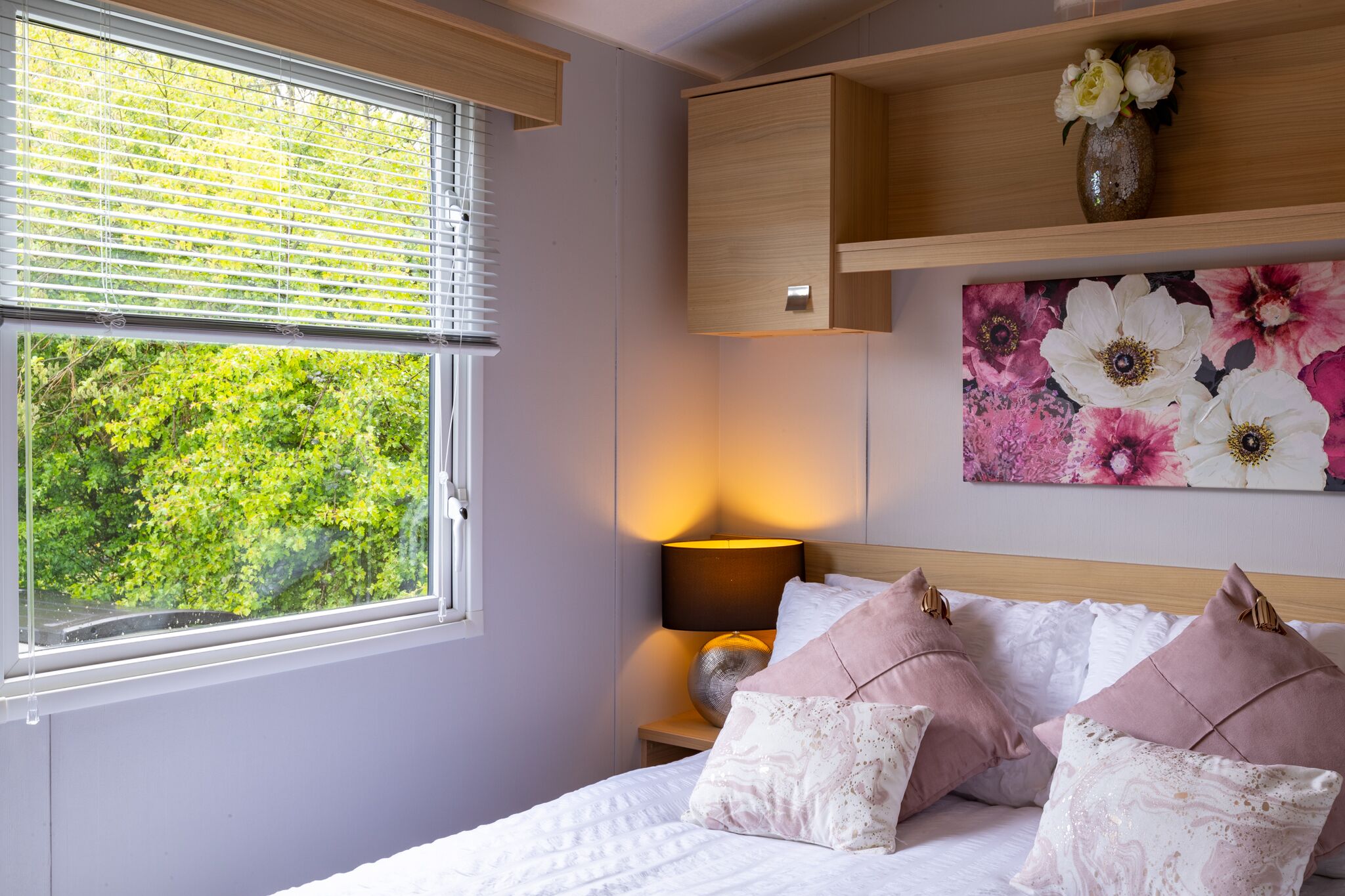Une chambre à coucher comprenant un lit, une table de chevet, une lampe, une fenêtre avec des stores, une étagère en bois ornée d’un vase et un tableau représentant des fleurs.