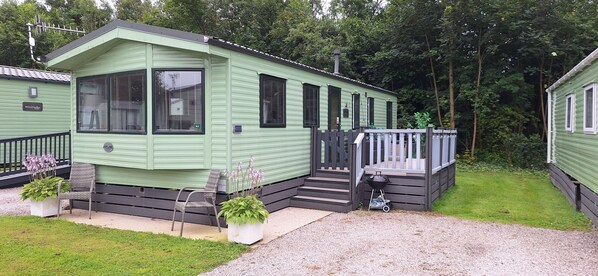 A green mobile home with a porch, a grill, and potted plants.