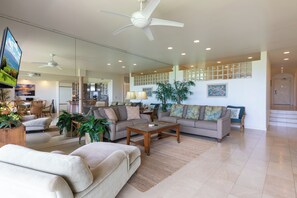 A spacious living room with a large sofa, a coffee table, and a TV. There are ceiling fans, a large window, and a staircase leading to another level.