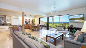 A spacious living area with a large window offering a view of the ocean, a dining area with a kitchen in the background, and a ceiling fan. - Ground-Floor Paradise:  Makena Surf F-110, Your Oceanfront Getaway! (Makena, Maui)