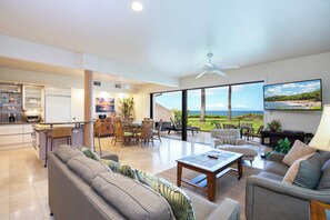 A spacious living area with a large window offering a view of the ocean, a dining area with a kitchen in the background, and a ceiling fan. - Ground-Floor Paradise:  Makena Surf F-110, Your Oceanfront Getaway! (Makena, Maui)