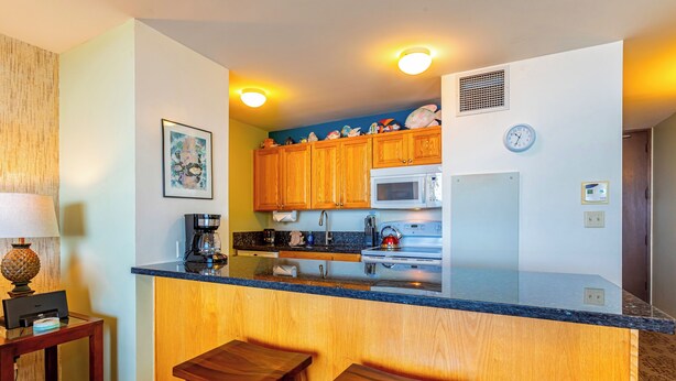 A compact kitchen with wooden cabinets, a microwave, and a sink. There are two wooden stools and a small table with a lamp.