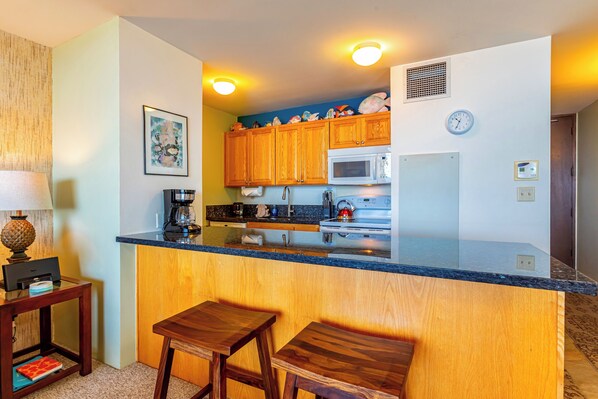 A compact kitchen with wooden cabinets, a microwave, and a sink. There are two wooden stools and a small table with a lamp.