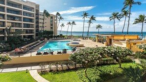 A hotel pool area with a view of the ocean, palm trees, and a building with multiple balconies.