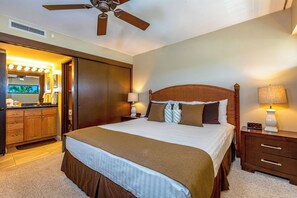 Ein Hotelzimmer mit einem großen Bett, einem Holzkopfstück, zwei Nachttischen mit Lampen, einem Deckenventilator und einem Badezimmer mit Waschtisch und Spiegel.