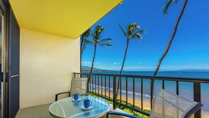 A balcony with a glass table, two chairs, and a view of the beach and ocean.