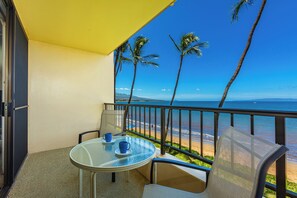 A balcony with a glass table, two chairs, and a view of the beach and ocean.