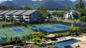 Un court de tennis entouré d’une végétation luxuriante et bordé d’une piscine, avec une vue sur les montagnes en arrière-plan.