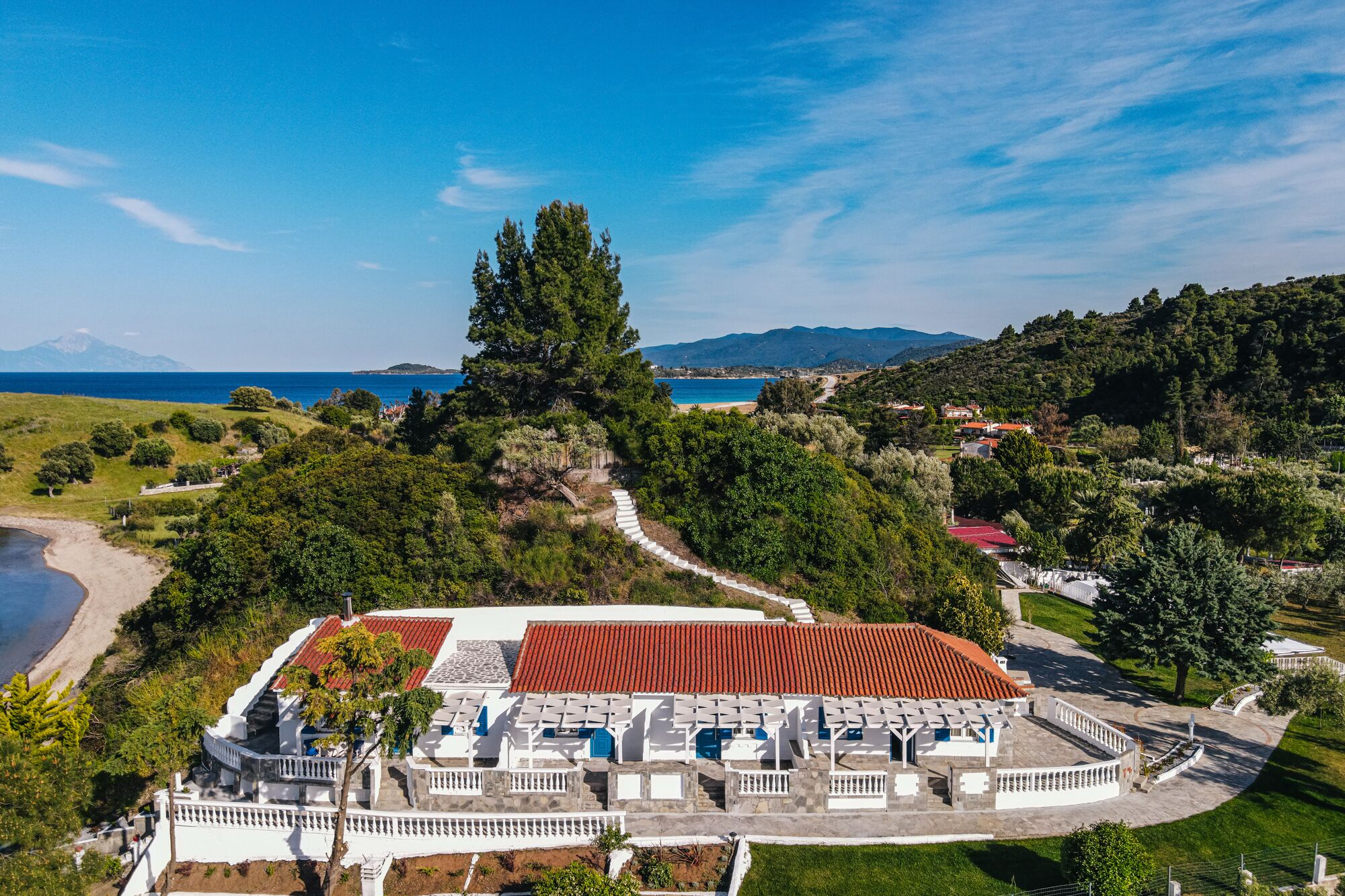 An aerial view of a coastal resort with a central building surrounded by lush greenery, a beach, and a mountainous backdrop.