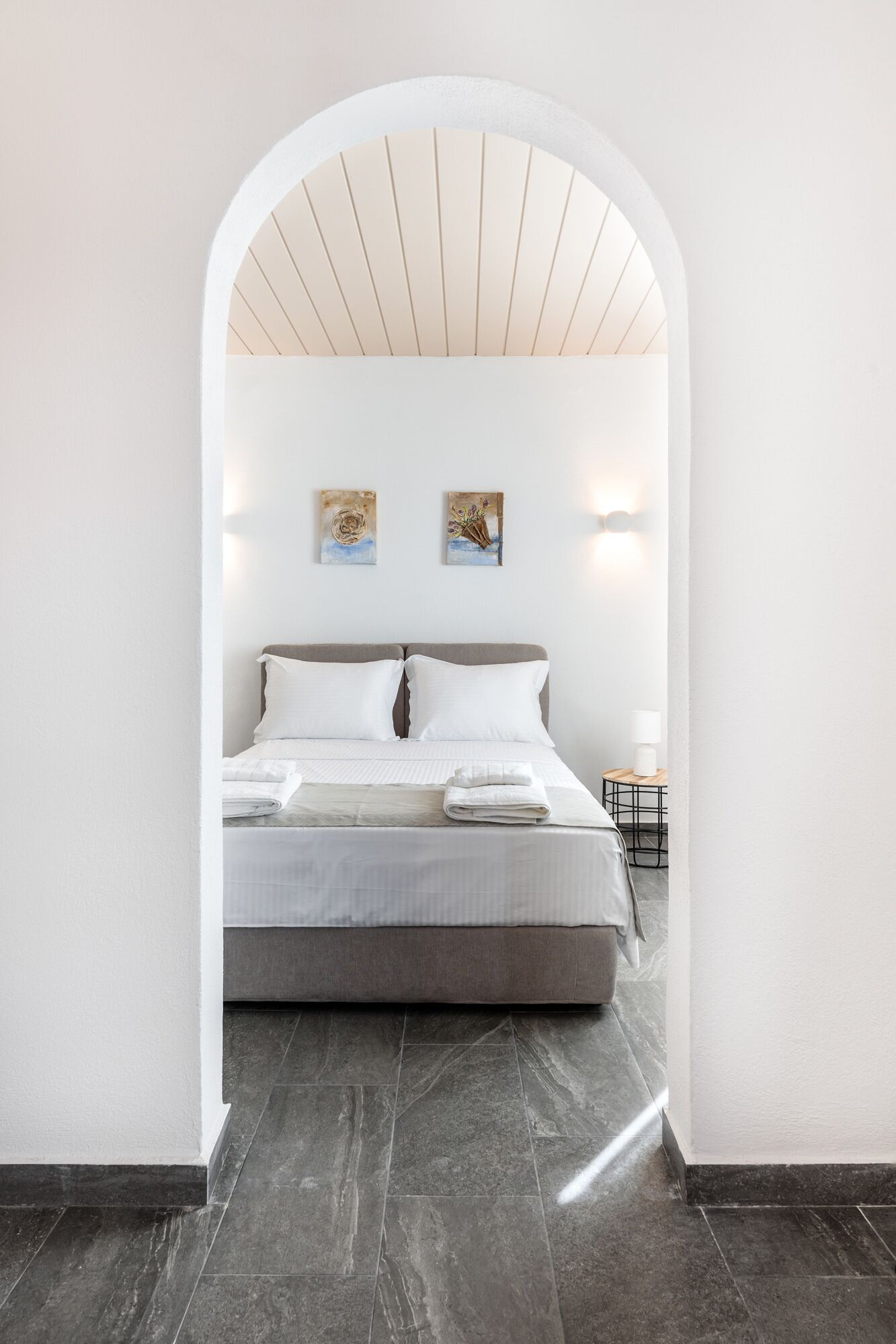 A modern bedroom with a bed, white walls, and a stone floor. There are two paintings on the wall and a small table with a lamp.