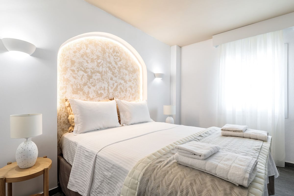 A hotel room with a large bed, a round headboard, a bedside table with a lamp, and a window with curtains.