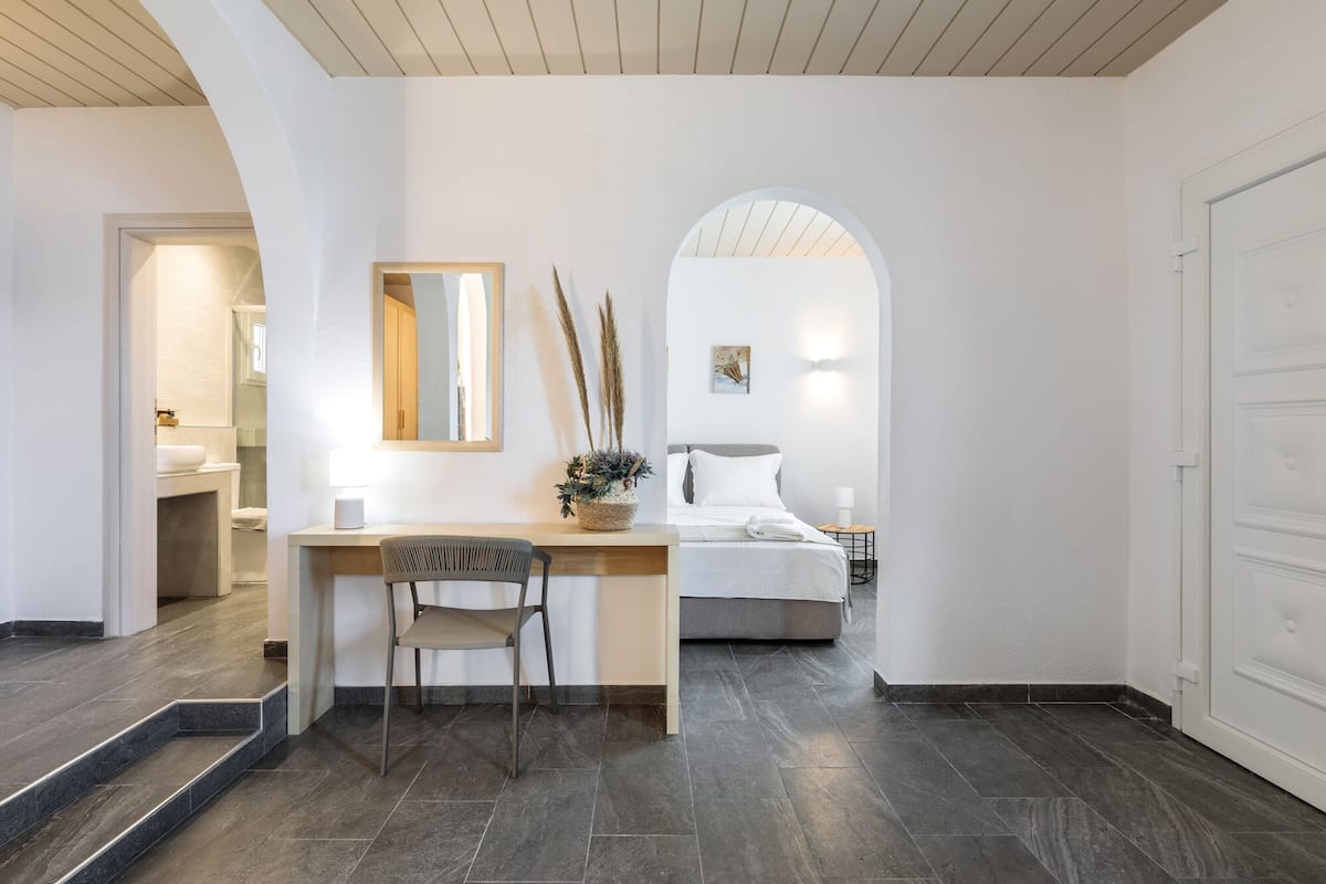 A modern hotel room with a wooden desk, a chair, a bed, and a bathroom visible through an archway.