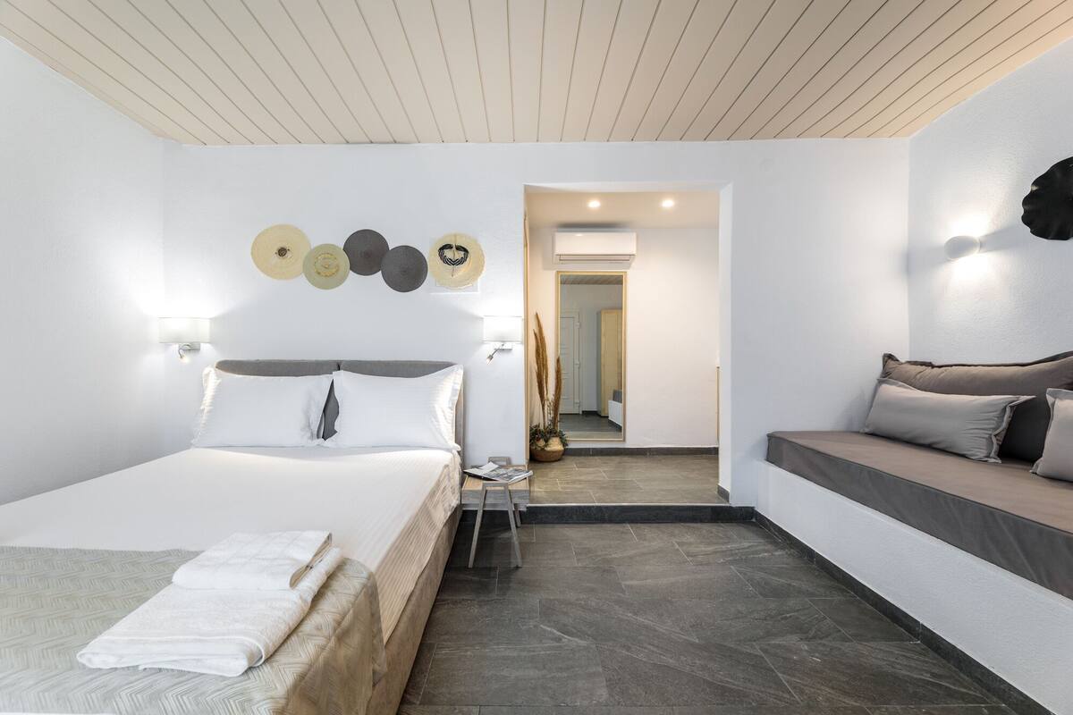 A modern hotel room with a bed, a sofa, and a small table. There is a hallway leading to another room.