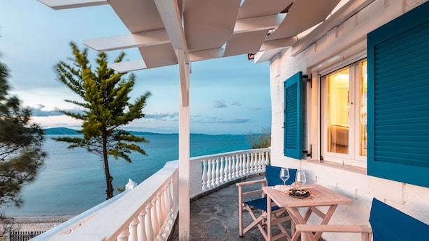 Un balcón con vista al mar, una mesa con sillas y una ventana con contraventanas azules.