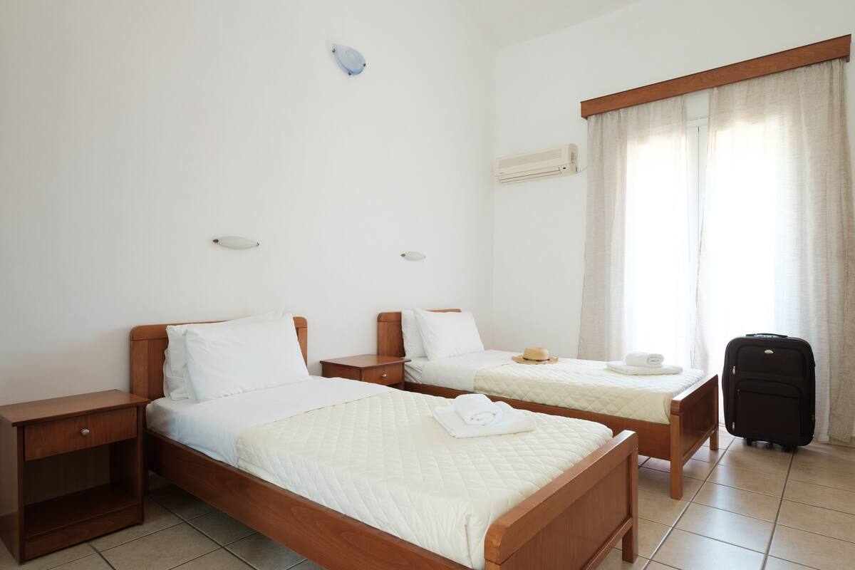 Una habitación de hotel con dos camas individuales, una mesita de noche de madera, una pequeña mesa y una maleta.