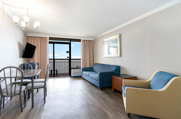 Habitación de hotel con una mesa de comedor, dos sofás, un televisor y un amplio ventanal con vistas al mar.