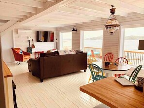 A cozy living room with a sofa, a dining table, and chairs, a guitar, and a view of the water. - Spencer Salt-Side Cottage (Twillingate) (Twillingate)