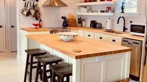 A kitchen with a central island, bar stools, hanging pots, and a window. - Spencer Salt-Side Cottage (Twillingate) (Twillingate)