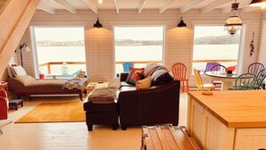 A cozy loft-style room with a wooden floor, a sofa, a dining table, and a view of the water. - Spencer Salt-Side Cottage (Twillingate) (Twillingate)