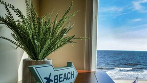 Una pianta in vaso e un cartello direzionale con la scritta 'BEACH', posati su un tavolo di legno vicino a una finestra con vista sull'oceano.
