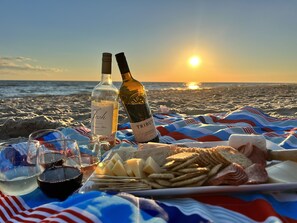 A beach picnic setup with two wine bottles, glasses, and a plate of snacks under a blanket. - Beach Please Oceanfront (Oak Island)