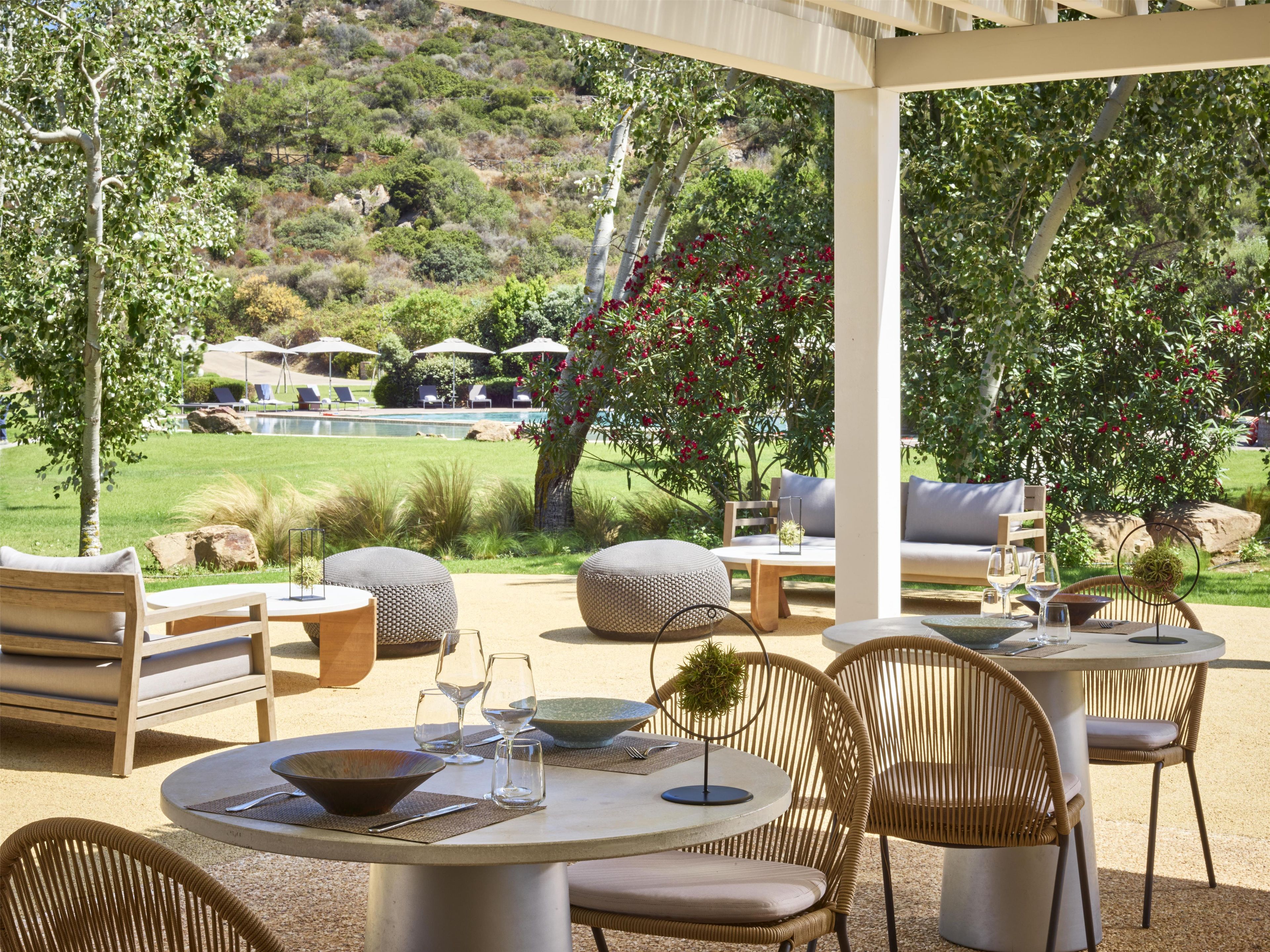 An outdoor patio with round tables and wicker chairs, overlooking a pool and lush greenery.
