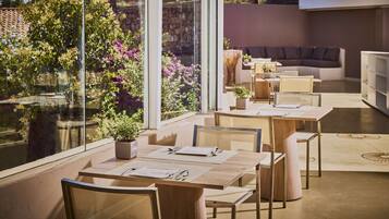 A modern dining area with glass walls, a wooden table set for two, and a cozy seating area with a sofa and chairs.