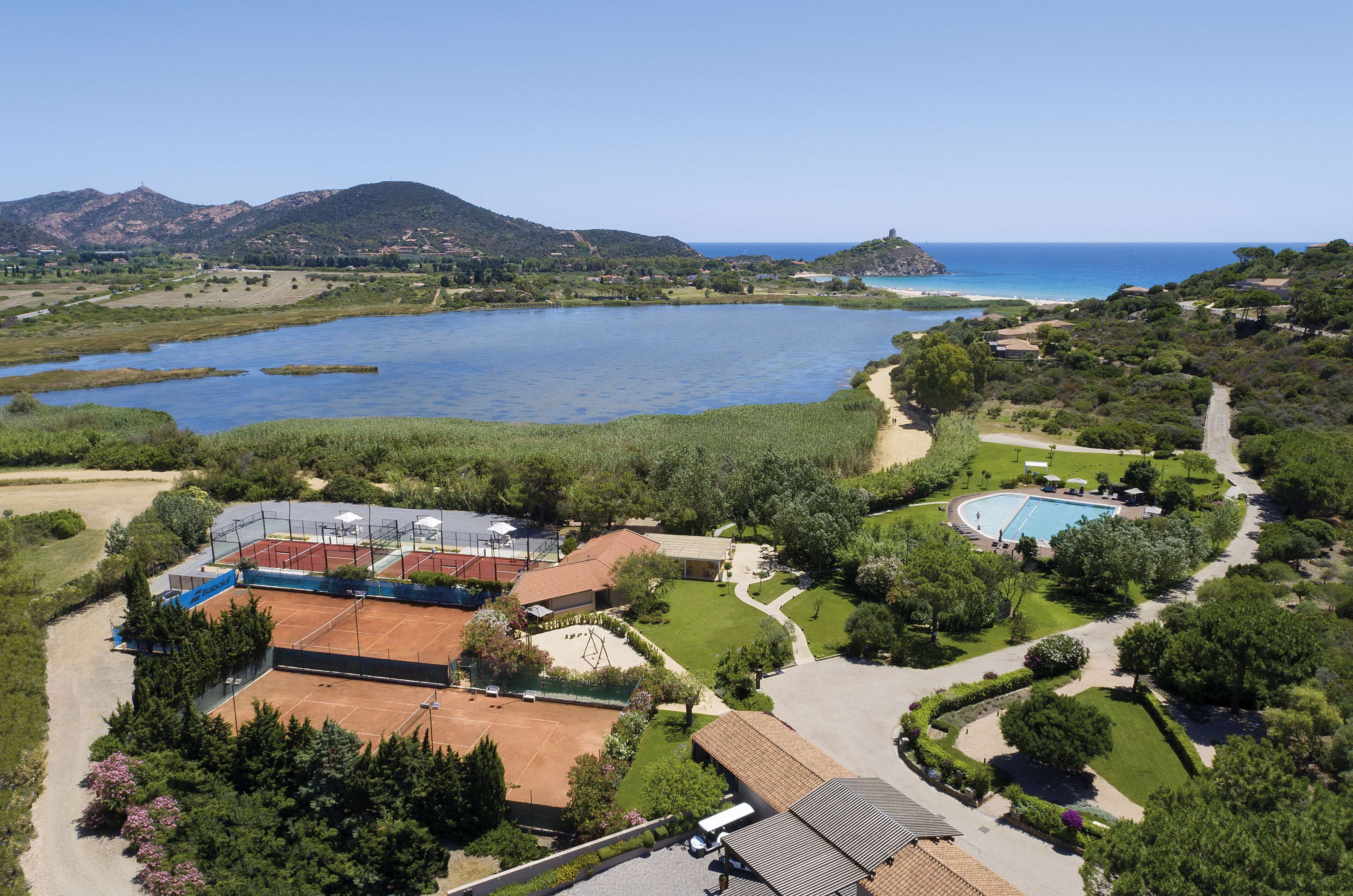 An aerial view of a resort with a tennis court, swimming pool, and a scenic lake with mountains in the background.