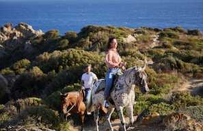 Dos personas montando a caballo por un sendero costero con terreno rocoso y vegetación.