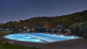 Ein großer Außenpool mit mehreren Liegebereichen und Sonnenschirmen, umgeben von einem angelegten Garten und einer sanften Hügelkulisse im Hintergrund.