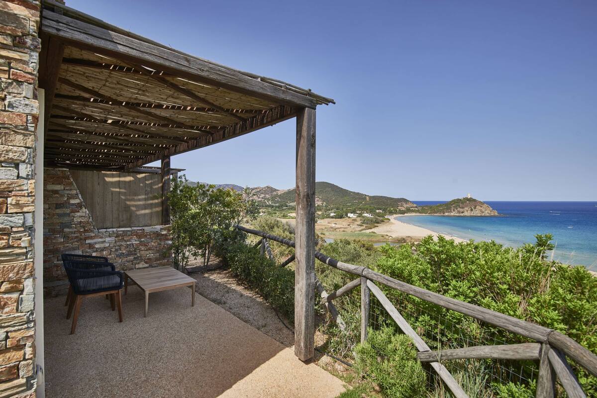 Ein Gebäude aus Stein mit einer Holzpergola und einer Sitzgelegenheit mit Blick auf einen Strand.