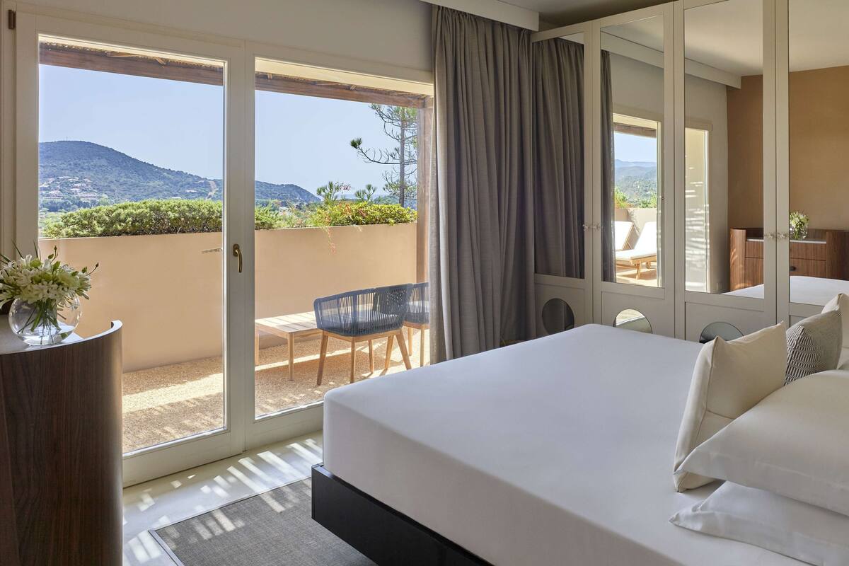 Ein Hotelzimmer mit einem großen Bett, einer Glasschiebetür, die auf einen Balkon mit Blick auf die Hügel führt, einer kleinen Sitzecke mit Stühlen und einer Holzkommode.