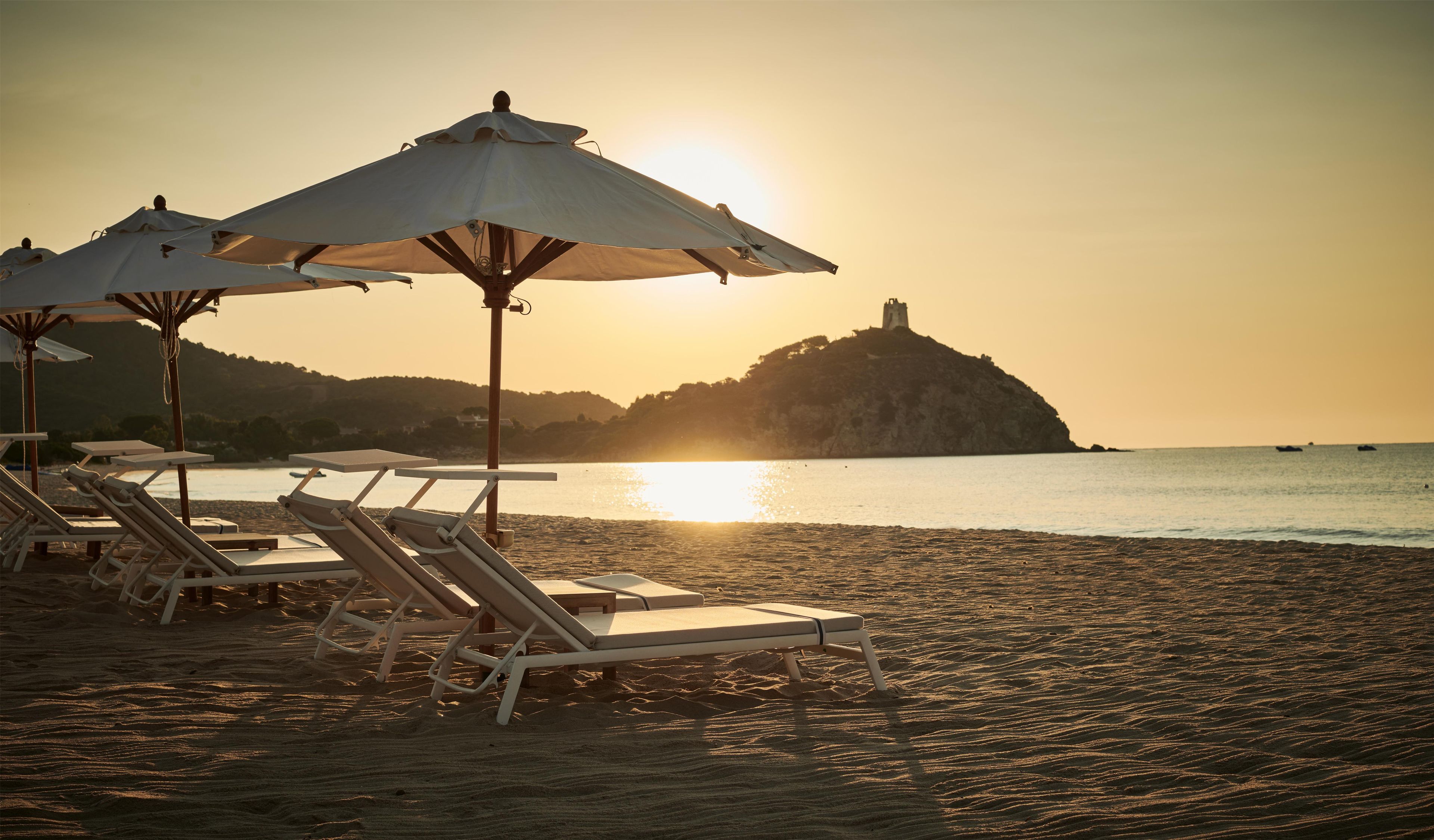 A beach with sun loungers and umbrellas, a clear sky, and a distant hill with a structure.