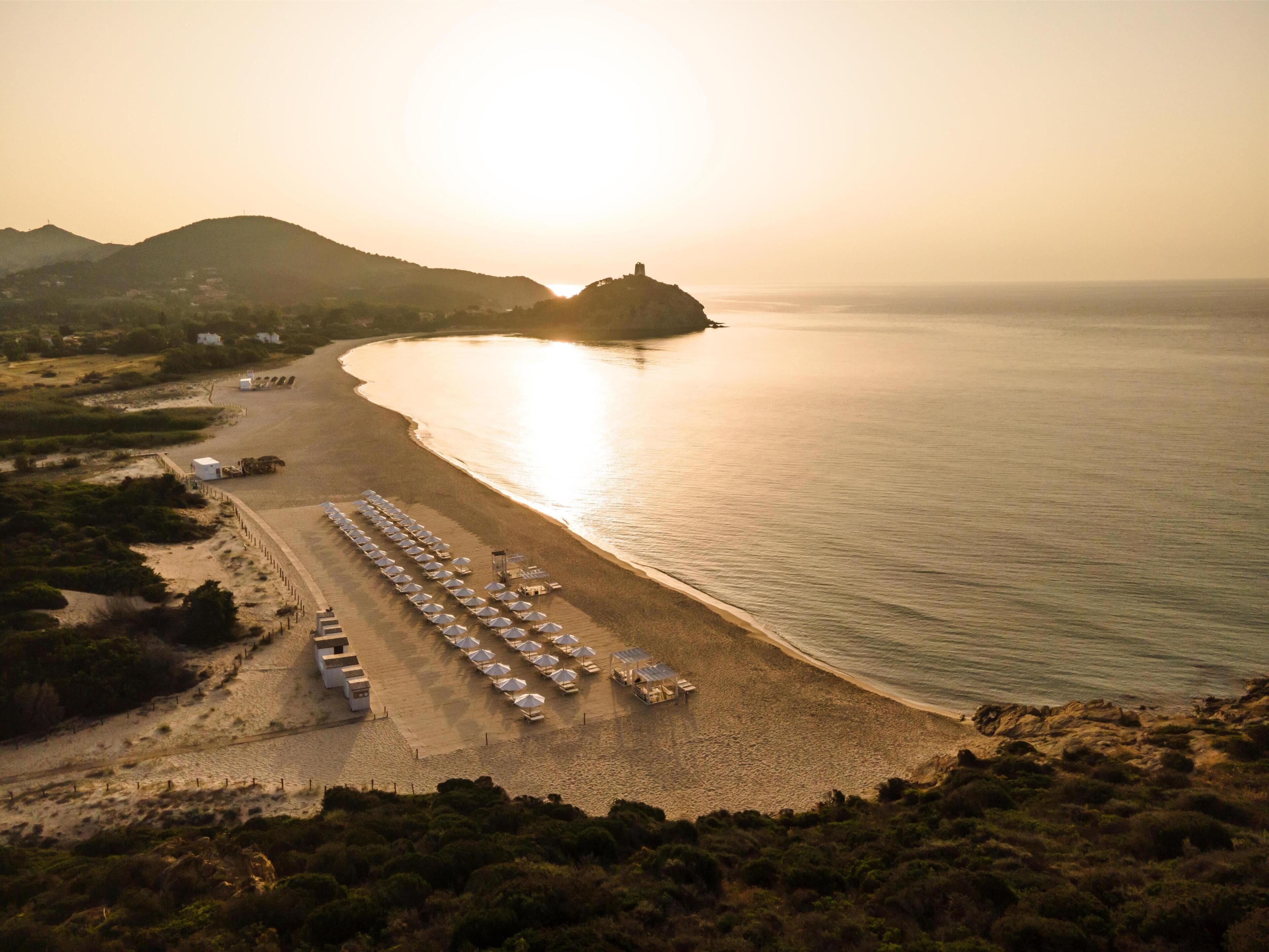 An aerial view of a beach with rows of umbrellas and beach chairs, a small building, and a distant hill.