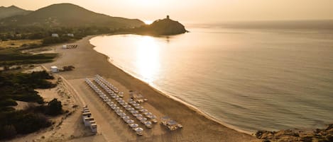 An aerial view of a beach with rows of umbrellas and beach chairs, a small building, and a distant hill.