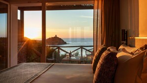 Ein Schlafzimmer mit großem Fenster und Blick auf die Küste, ein Bett mit Kissen sowie ein kleiner Tisch mit Lampe.