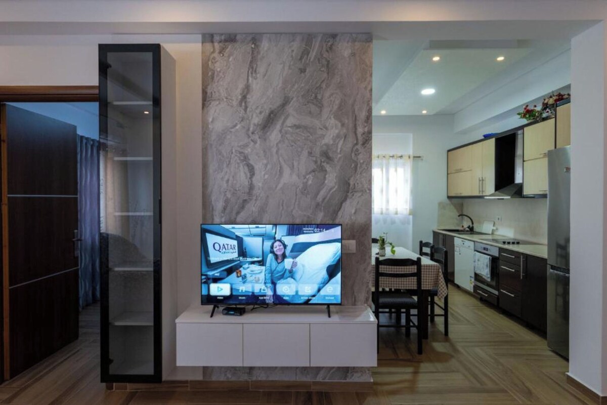 A modern living room with a flat-screen TV on a white console, a marble accent wall, a dining area with a checkered tablecloth, and a kitchen with dark cabinetry and a stainless steel refrigerator.