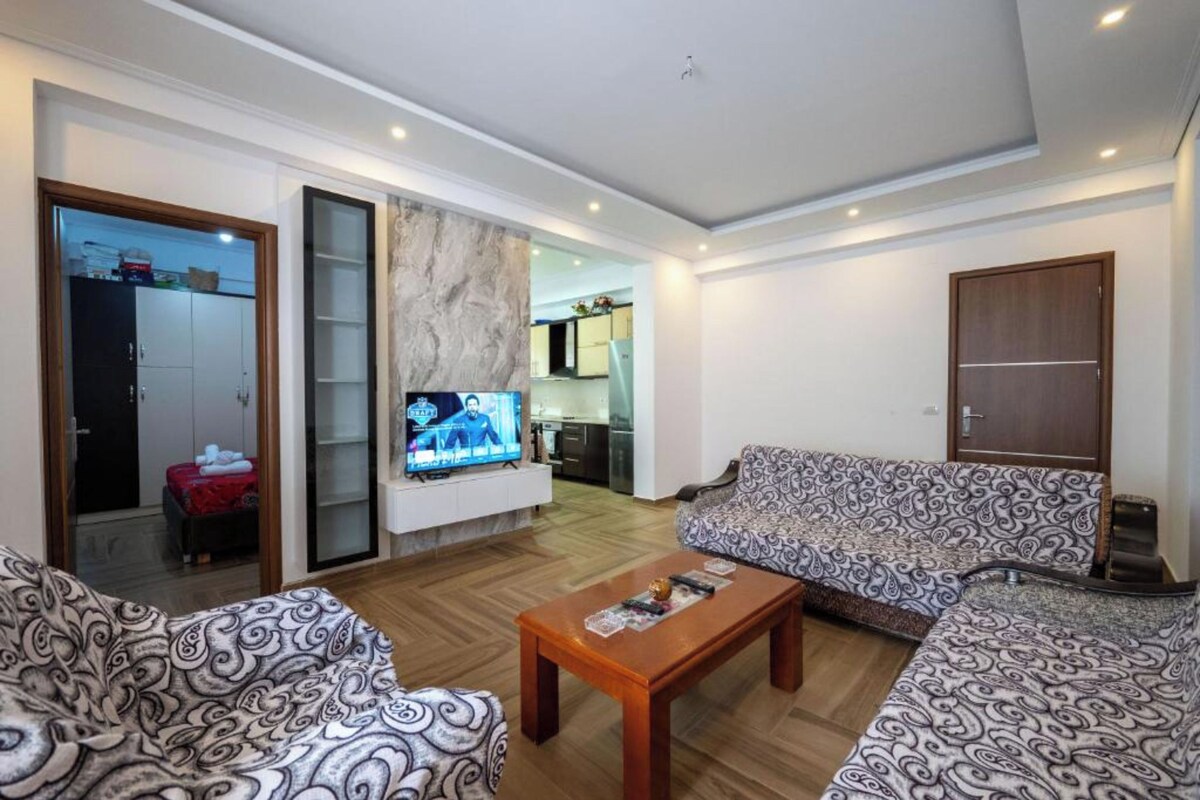 A living room with patterned sofas, a wooden coffee table, a TV, and a sliding glass door leading to a kitchen.