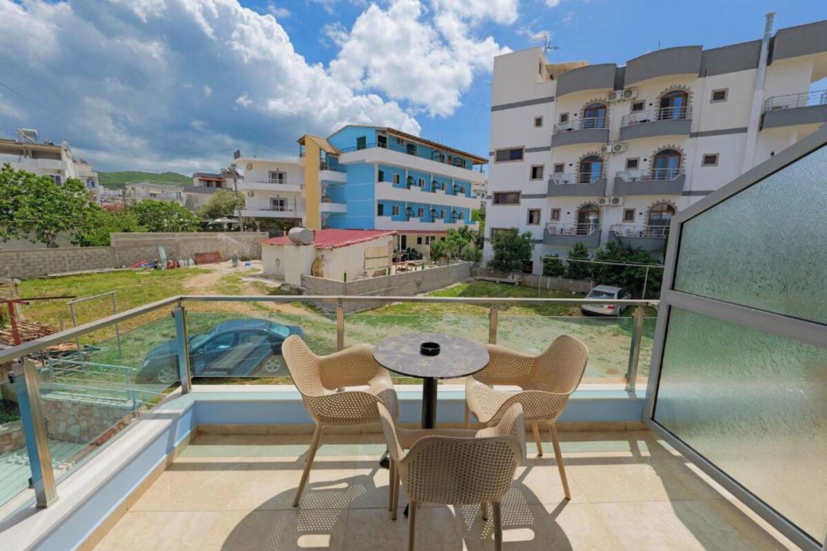 A balcony with wicker chairs and a table, overlooking a residential area with multi-story buildings and a parked car.