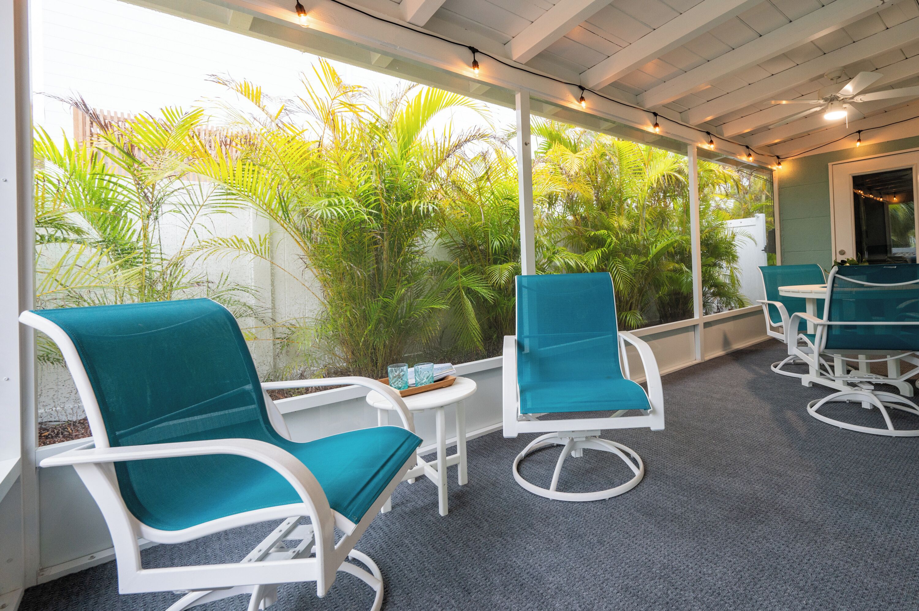 Un espace extérieur aménagé, abrité et doté de chaises et de tables de couleur turquoise, entouré d’une végétation luxuriante.